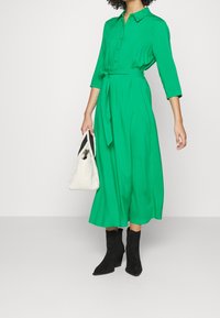 Grünes langärmeliges Kleid mit Kragen, tailliertem Bund und fließendem Rock. Schwarze Knöchelstiefel und eine weiße strukturierte Handtasche runden den Look ab.