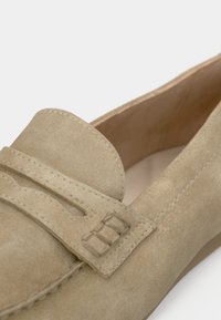 Mocassins en daim beige avec un détail cousu et une lanière décorative. Bout arrondi et doublure intérieure lisse. Semelle plate pour un port facile.
