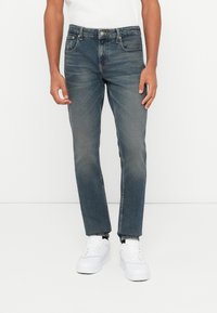 Mees, kellel on seljas kulunud sinised teksapüksid ja valged spordijalatsid, seisab ühetoalise valge tausta ees.