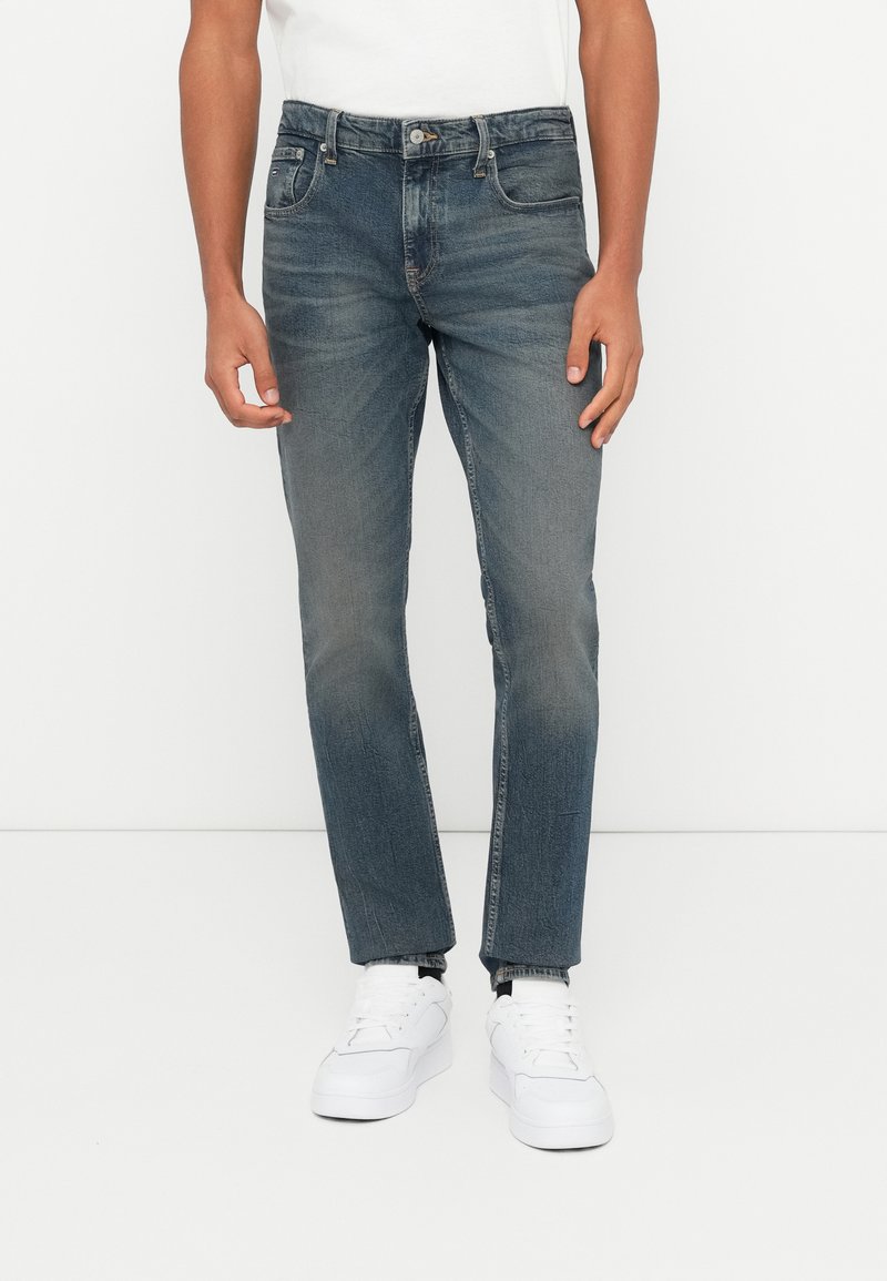 Mees, kellel on seljas kulunud sinised teksapüksid ja valged spordijalatsid, seisab ühetoalise valge tausta ees.