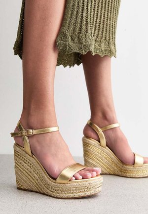 Gouden wig-sandalen met een gevlochten jute platform, voorzien van glanzende metalen bandjes en een gesp, gecombineerd met een gemarmerde groene gebreide jurk.