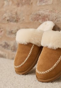 Pantofole in suede marrone con interno in pelliccia sintetica color crema, cuciture bianche e una texture morbida, posizionate su uno sfondo neutro.