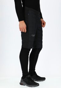 Zwarte geïsoleerde shorts met een doorgestikt ontwerp, gedragen over strakke zwarte leggings. Bevat een logo en een comfortabele elastische tailleband.