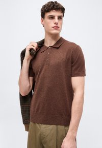 Νέος άνδρας που φοράει καφέ κοντομάνικο πόλο, κρατώντας ένα καρό σακάκι πάνω στον ώμο του, στέκεται μπροστά σε απλό φόντο.