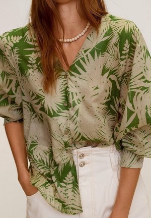 Frau trägt eine grün-beige Bluse mit tropischem Blattmuster und hochgekrempelten Ärmeln, weiße hochtaillierte Hose und eine Perlenkette.