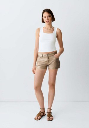 Frau steht, trägt ein weißes ärmelloses Oberteil, beige Shorts und braune mit Nieten besetzte Sandalen vor einem schlichten hellen Hintergrund.