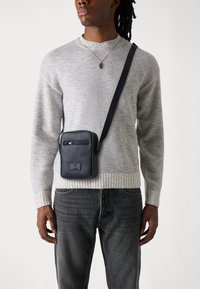 Borsa a tracolla in pelle nera con finitura texturizzata, tasca frontale con zip e tracolla regolabile, indossata sopra un maglione grigio texturizzato.
