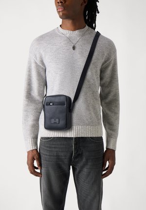Borsa a tracolla in pelle nera con finitura texturizzata, tasca frontale con zip e tracolla regolabile, indossata sopra un maglione grigio texturizzato.