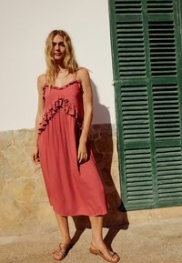 Frambozenroze midi-jurk met gerimpelde accenten, dunne bandjes en een losse pasvorm, gecombineerd met platte sandalen tegen een gestructureerde stenen achtergrond.