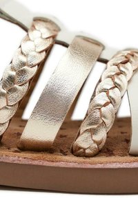 Tiras metálicas trenzadas en dorado con acabado suave sobre una base clara, colocadas sobre una plantilla texturizada color beige con sutiles patrones y una suela plana.