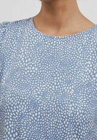 Blå blus med spridda oregelbundna vita prickar och rund krage, bärs av en person.