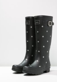 Botas de lluvia de goma negras con un patrón de estrellas en plata, un detalle de hebilla en el lateral y una suela texturizada para agarre.
