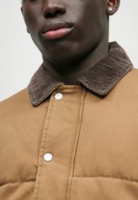 Geacă călduroasă de culoare maro, cu guler din corduroy ribat și nasturi metalici argintii. Materialul are un aspect texturat și durabil.