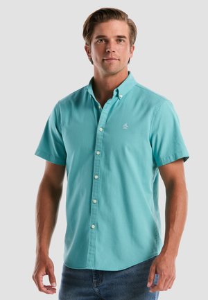 Man in een turkooizen korte mouwen overhemd met kleine logo op de borst, staand tegen een effen lichte achtergrond.