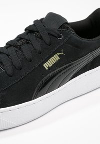 Baskets en daim noir Puma avec lacets noirs, bande latérale noire brillante, logo Puma doré et semelle blanche texturée.