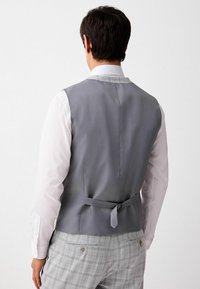 Gray fabric vest with a smooth texture, featuring a back tie detail, worn over a white shirt with long sleeves, paired with checked trousers.