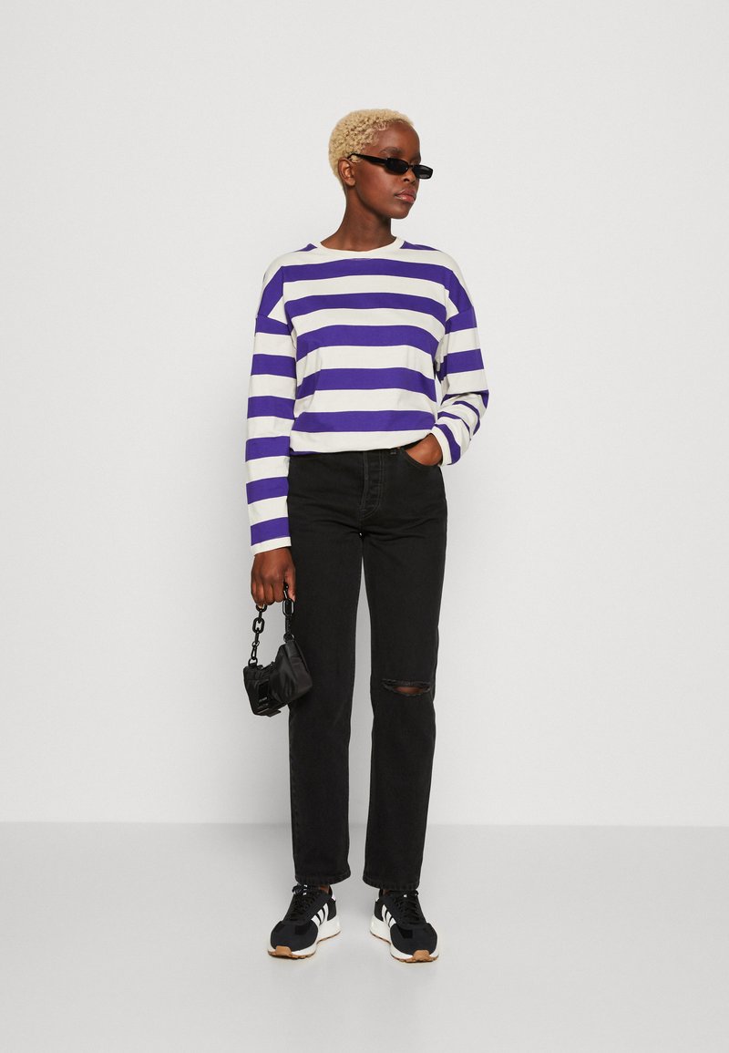 Camisa de rayas de manga larga en blanco y morado, vaqueros negros rasgados, zapatillas negras con rayas blancas y un pequeño bolso negro.