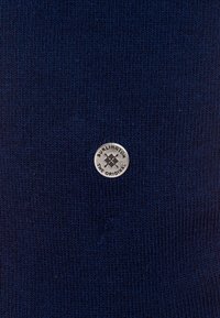 Granatowy dzianinowy materiał z prążkowaną teksturą. Posiada okrągłe, srebrne logo z napisem "BURLINGTON THE ORIGINAL" na środku.