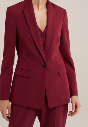 Femme portant un tailleur rouge profond sur mesure avec un blazer boutonné à l'avant et un pantalon assorti sur un fond neutre.