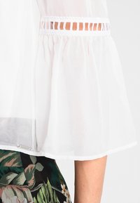 Weißes, transparentes Blusenshirt mit ausgestellten Ärmeln und Spitzen-Ausschnitten. Der Saum ist gerade, mit einem Etikett am unteren Ende. Kombiniert mit einem Rock mit floralem Muster.