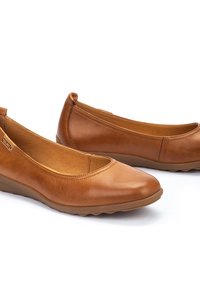 Mocasines de cuero marrón con punta redonda y suela baja tipo cuña. Textura suave con un diseño minimalista. Inserciones elásticas laterales para facilitar el uso.