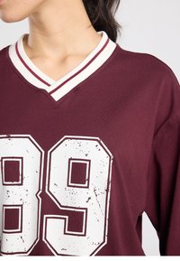 Chemise oversize couleur bordeaux avec col en V et col à rayures blanches. Présente de grands chiffres blancs usés "89" à l'avant. Texture de tissu doux.