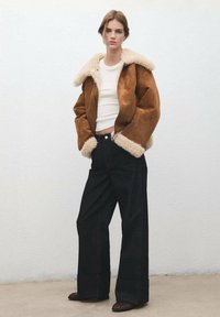 Bruine suède jas met een crème kleurige shearling kraag, wit aansluitend overhemd en zwarte wijde broekspijpen. Gestructureerd materiaal en een losse pasvorm.