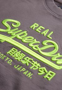 Grijze sweatshirt met vet, verhoogde neongele borduursel. Het ontwerp bevat "SuperDry" met Aziatische karakters en "Tokyo, Japan."