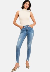 Kitsad kõrge vöökoht sinised teksad, millel on kerge kulumise efekt; koos beeži ilma varrukateta kõrge kaelusega pluusiga ja kaunistatud kõrge kontsaga kingadega.