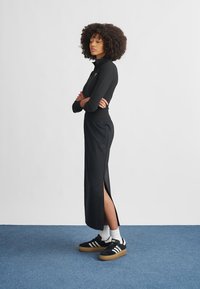 Svart langermet topp med høy krage og tettsittende design, kombinert med svart maxiskjørt med sidelommer. Svart-hvite sneakers med gummisåler.