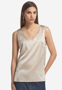 Satin, ärmlös beige topp med V-ringning. Har en avslappnad passform och subtil lyster, kombinerad med blå denimbyxor.