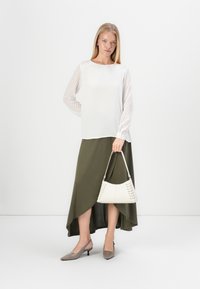 Weiße Bluse mit gemusterten Ärmeln, olivgrüner asymmetrischer Rock, graue spitze Pumps und weiße Handtasche mit Schnürdetails.