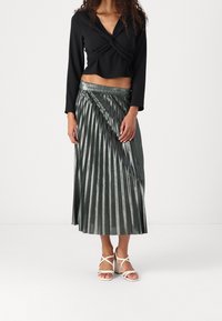 Top nero a maniche lunghe, corto, con davanti annodato; gonna midi plissettata verde con finitura metallizzata; sandali con zeppa bianchi e cinturino.