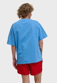 T-shirt en coton bleu clair avec une coupe décontractée, des manches courtes et un col rond. Porté avec un short rouge ; la vue de dos montre une texture lisse.