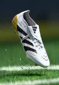 White soccer cleat with black and gold accents, textured synthetic upper, six cleats on the sole, floating above grass with debris scattering.