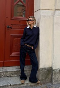 Marineblaue Langarm-Polo mit weißen Kragen, kombiniert mit weiten, dunklen Denim-Jeans und braunen Slippern, die vor einer roten Tür stehen.