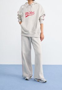 Světlé šedé mikina Nike s červeným logem, klokánkou a šňůrkami na stažení. Doplněné o odpovídající tepláky s červenými bočními pruhy.