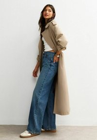 Mujer con un abrigo trench beige, camiseta blanca sin mangas, jeans de pierna ancha azul y zapatillas blancas, de pie en un ángulo contra una pared blanca.