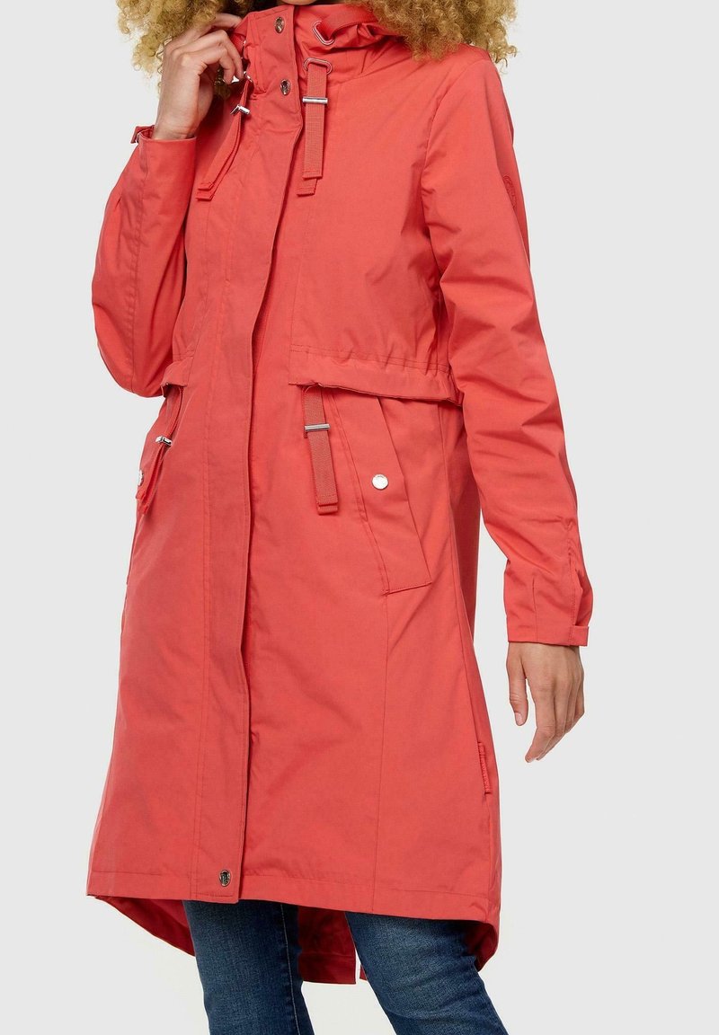 Kobieta w długiej koralowej kurtce z guzikami i detalami w postaci pasków, stoi z jedną ręką przy twarzy, a drugą swobodnie zwisającą wzdłuż ciała.