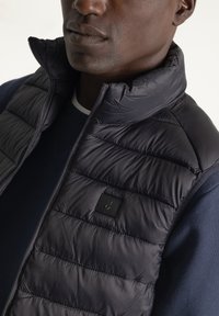 Chaleco de plumas azul marino con patrón de acolchado vertical, cuello alto y cierre de cremallera. Presenta una pequeña etiqueta de marca negra en el pecho.