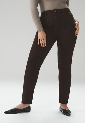 Vrouw draagt nauwsluitende donkerbruine broek, lichtbruine longsleeve top, zwarte puntige platte schoenen en ringen aan linkerhand, staand op effen achtergrond.