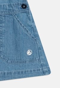 Sv�ětlá modrá denímová látka s špičatou kapsou, černým knoflíkovým detailem a bílou vyšitou logem plachetnice poblíž kapsy.