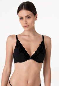 Donna dai capelli scuri che indossa un reggiseno nero con ricami floreali, posando davanti a uno sfondo bianco uniforme.