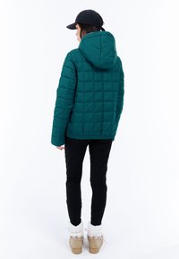 Persoon in een teal gekwilt jack met capuchon, zwarte broek, tan winterlaarzen met witte bontvoering en een zwarte pet, staande met de rug naar de camera.