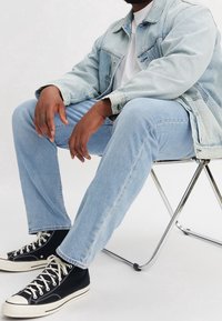 Man sitter på en metallstol iförd en ljus denimjacka, ljusa jeans, vit t-shirt och svarta högskor med vita snören.