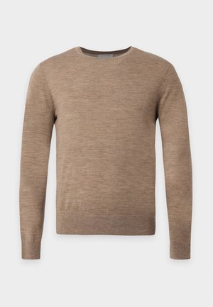 Uomo che indossa un maglione marrone a maniche lunghe, jeans blu scuro e sneakers bianche, in piedi con una mano in tasca su sfondo semplice.
