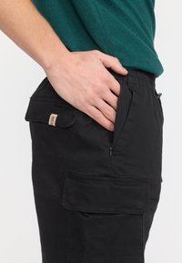 Main insérée dans la poche latérale d'un pantalon cargo noir porté avec une chemise verte, montrant les détails de la fermeture éclair et de la poche à rabat.