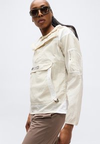 Άτομο φορώντας σκούρα γυαλιά ηλίου, μπεζ μπουφάν Columbia με μπροστινή τσέπη και τσέπη από δίχτυ στο μανίκι, καθώς και ανοιχτό καφέ παντελόνι, σε απλό φόντο.