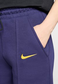 Lilla joggebukser laget av myk stoff har en gul Nike-logo på venstre lår, elastisk midjebånd og sidelommer.