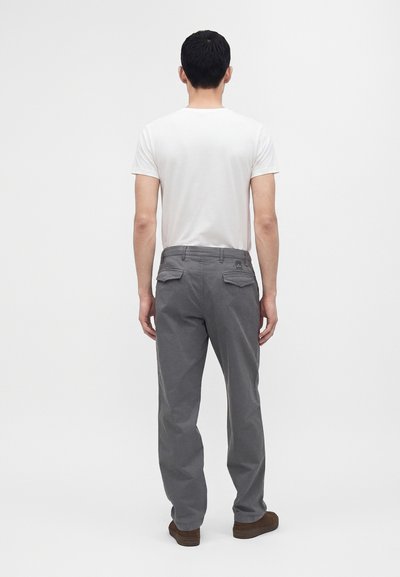 Homme debout, dos tourné, portant une chemise blanche à manches courtes, un pantalon gris avec des poches à l'arrière et des chaussures marron sur un fond uni.